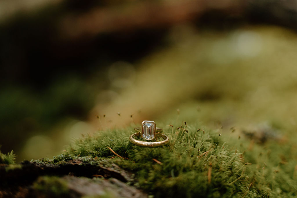 Emerald cut engagement ring and gold wedding band placed on a moss-covered log during an adventurous elopement in Montana