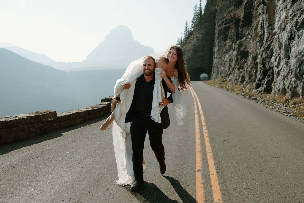 A groom lifts his bride over his shoulders as they frolic along Going to the Sun Road in Glacier National Park